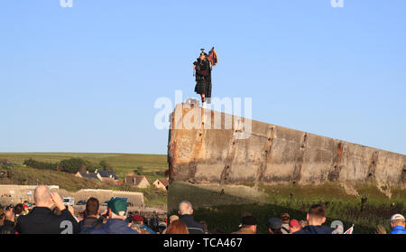 Ein einsamer Piper spielt auf den Maulbeerbäumen Hafen von Arromanches in der Normandie, im Norden Frankreichs, als britische Armee den genauen Moment Mark der erste britische Soldat auf Gold Beach vor 75 Jahren landete. Stockfoto