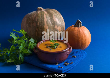 Kürbis Suppe in eine Schüssel geben und mit Petersilie, Kürbiskerne und zwei Kürbisse auf dem blauen hellen Hintergrund diente, horizontale Ausrichtung Stockfoto