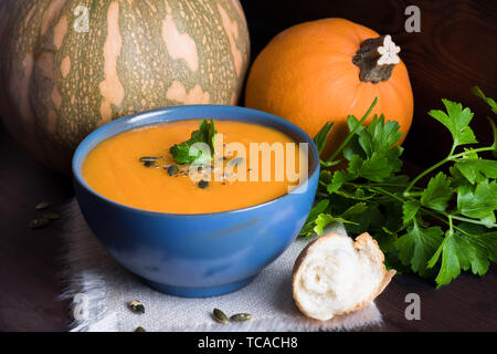 Kürbis vegane Suppe in eine blaue Schale serviert mit Petersilie, Olivenöl und Kürbiskerne und Brot auf einem dunklen Hintergrund Stockfoto