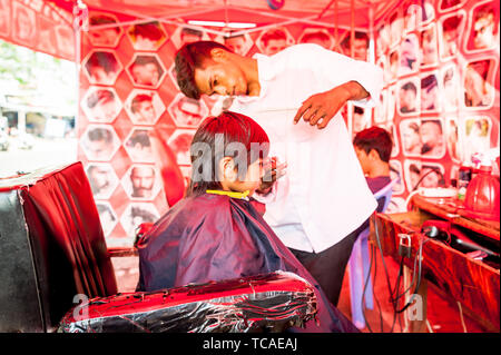 Ein schüchterner kleiner kambodianischer Junge bekommt seine Haare in einem typischen Barbershop im Freien in Phnom Penh, Kambodscha geschnitten. Stockfoto