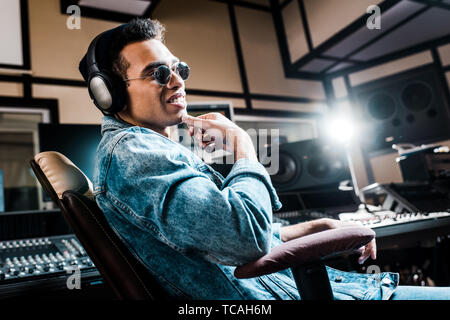 Lächelnd Mixed Race Sound Hersteller in Kopfhörer und Sonnenbrille sitzt im Recording Studio Stockfoto
