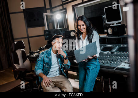 Lächelnd hübscher Ton Produzent mit Laptop in der Nähe von gemischten Rennen Freund in Recording Studio Stockfoto