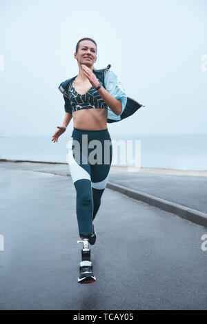 Nicht ohne Sport leben. Vertikale Foto von stark behinderte Frau in Sportkleidung läuft im Freien in der Nähe des Meeres und lächelnd. Motivation. Sport Konzept. Behinderte Sportler Stockfoto