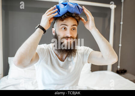 Portrait eines bärtigen Mannes mit Eisbeutel auf dem Kopf, mit Kater im Laufe des Vormittags auf dem Bett Stockfoto