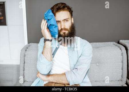 Bärtiger Mann mit Kopfschmerzen Anwendung Eisbeutel, während auf der Couch zu Hause sitzen Stockfoto