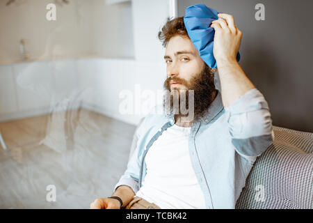 Bärtiger Mann mit Kopfschmerzen Anwendung Eisbeutel, während auf der Couch zu Hause sitzen Stockfoto