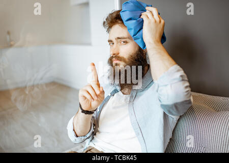 Bärtiger Mann mit Kopfschmerzen Anwendung Eisbeutel, während auf der Couch zu Hause sitzen Stockfoto