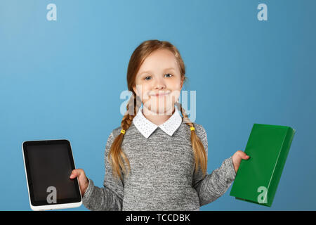 Fröhliche kleine Mädchen auf einem blauen Hintergrund. Das Kind hält ein Buch und eine Tablette. Der Begriff der Bildung, Schule, Auswahl, Vergleich. Stockfoto