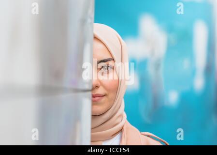 Schöne muslimische Frau in Kopftuch und modernen Kleidung versteckt die Hälfte Gesicht an der Wand mit blauen Hintergrund.