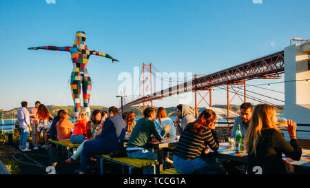 Lissabon, Portugal - Juni 5, 2019: Blick auf den 25. April Brücke mit Personen sitzen auf der Terrasse der Bar bei LX-Fabrik in Alcantara Stockfoto