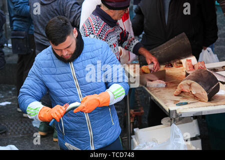 Catania, Italien - Apr 10 2019: Junge fischer Skalierung und Ausnehmen Fische auf dem berühmten Sizilianischen Fischmarkt. Herausziehen der Roe und Innereien ist ein notwendiger Teil der Fischverarbeitung. Touristische Attraktion. Stockfoto