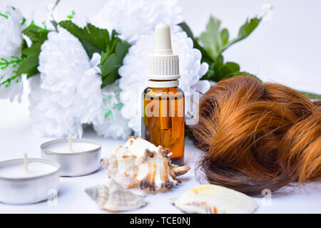 Arganöl Flasche mit, braune Haar. Das Glas hinter weißen Blüten, davor Muscheln und kleinen Teelicht Duftkerzen in Metallrahmen Stockfoto