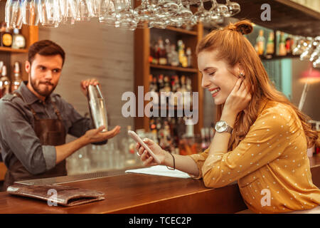 Mit Blick auf das Telefon. Glücklich lächelnde strahlende Langhaarigen bezaubernde Verführerische junge Frau mit Blick auf das Telefon in den Händen, während ein stattlicher attraktive adul Stockfoto