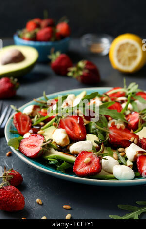 Gesunder Salat mit Erdbeeren, Avocado, Rucola und Mozzarella, mit Olivenöl und Balsamico Dressing in einer Platte auf einem dunklen Hintergrund entfernt gekleidet Stockfoto