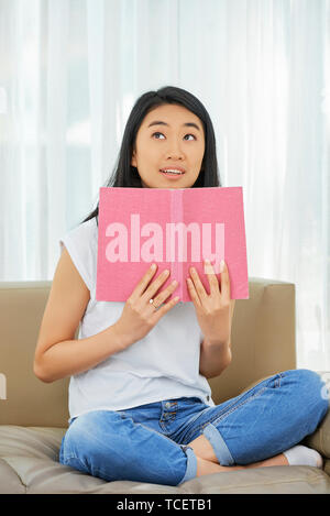 Portrait von positiven asiatischen Mädchen sitzt auf einem Sofa mit geöffnetem Buch und versuchen zu merken Material für Schule Stockfoto
