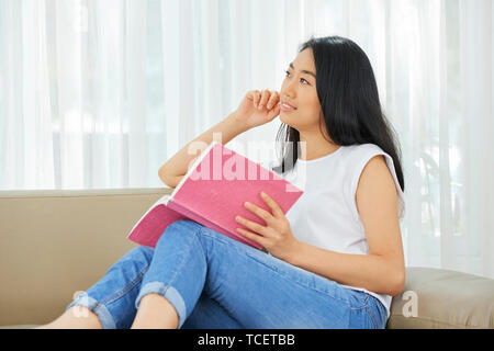 Ziemlich Vietnamesischen junges Mädchen auf einem Sofa mit geöffnetem Buch sitzen und träumen von etwas Stockfoto