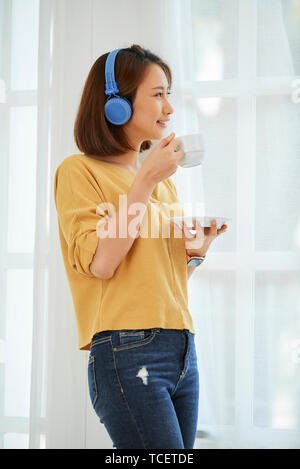 Hübsche junge asiatische Frau trinkt Kaffee am Morgen und Suchen durch große Fenster in Ihrem Zimmer Stockfoto
