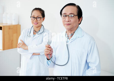 Portrait von positiven Vietnamesischen Allgemeinmediziner in weißen Laborkitteln Stethoskope Holding Stockfoto