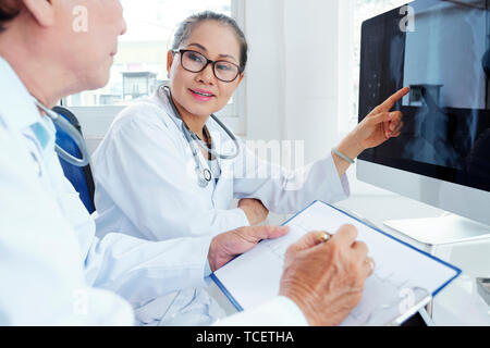 Lächelnd ziemlich vietnamesischen Arzt diskutieren Chest X-Ray auf dem Computerbildschirm mit erfahrenen Kardiologen Stockfoto