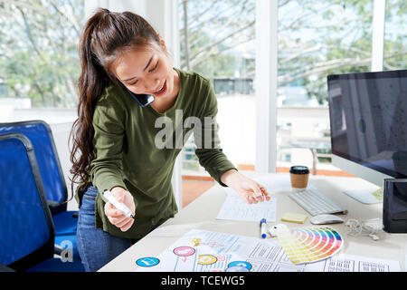 Fröhlicher junger asiatischer UI-Designer am Telefon sprechen mit Client und Korrekturen im Projekt Stockfoto