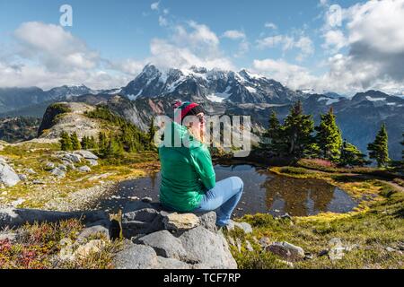 Weibliche Wanderer auf einem Felsen an einem kleinen Bergsee, Blick von der Tischplatte Berg Mt. Mit Schnee und Gletscher, Mt Shuksan. Baker-Snoqualmie Nation Stockfoto