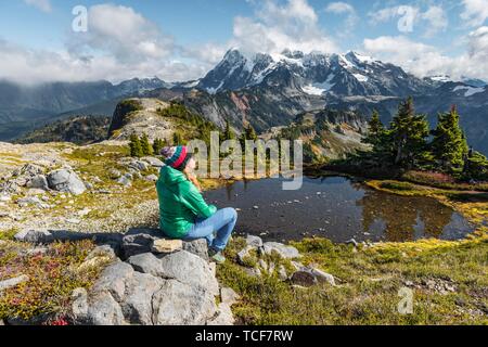 Weibliche Wanderer auf einem Felsen an einem kleinen Bergsee, Blick von der Tischplatte Berg Mt. Mit Schnee und Gletscher, Mt Shuksan. Baker-Snoqualmie Nation Stockfoto