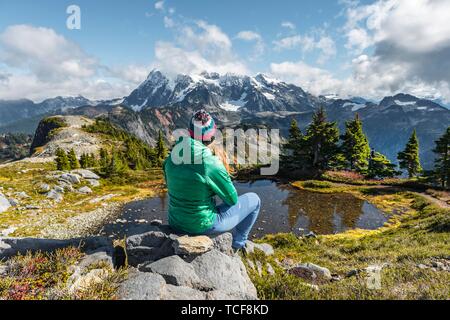 Weibliche Wanderer auf einem Felsen an einem kleinen Bergsee, Blick von der Tischplatte Berg Mt. Mit Schnee und Gletscher, Mt Shuksan. Baker-Snoqualmie Nation Stockfoto