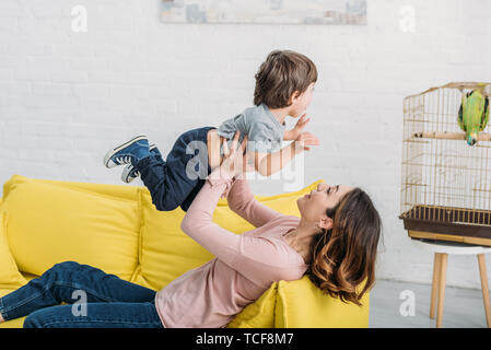 Glückliche junge Mutter mit entzückenden jungen Spaß auf gelben Sofa in der Nähe von Bird Cage mit Green Parrot Stockfoto