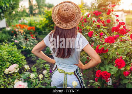 Junge Frau Sommer Garten bewundern. Gärtner in Schürze und Hut mit Blumen. Stolz auf das Ergebnis. Gartenarbeit Konzept Stockfoto