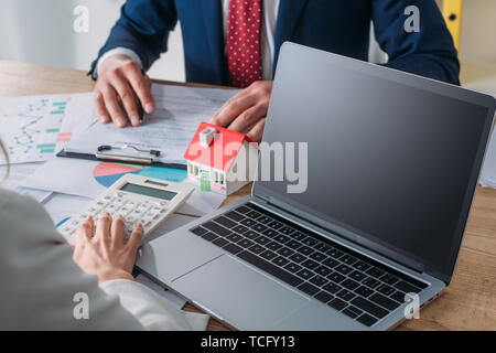 7/8 Schuß des Menschen studieren Darlehensvertrag und Geschäftsfrau mit Taschenrechner, während am Arbeitsplatz mit in der Nähe von Laptop, Haus Modell, Grafiken und ch Sitzung Stockfoto