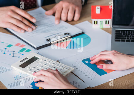 Teilansicht des Menschen studieren Darlehensvertrag und Geschäftsfrau mit Taschenrechner, während am Arbeitsplatz mit Haus Modell, Graphen und Diagramme sitzen Stockfoto