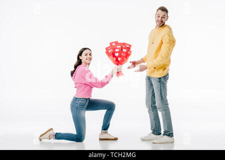 Hübsches Mädchen stehen auf Knie- und Holding Bouquet von roten Papier Cut Cards mit Herzen Symbole in der Nähe von lächelnden Freund auf weißem Hintergrund Stockfoto