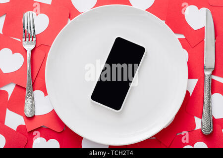 Draufsicht auf das Smartphone mit leeren Bildschirm auf weiße Platte in der Nähe der Messer und Gabel auf rotem Papier Cut Cards mit Herzen Symbole Stockfoto