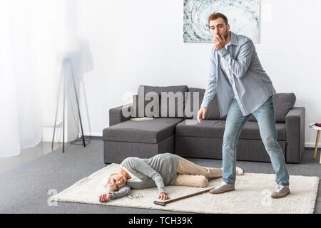 In voller Länge ansehen verunsicherter Mann stand in der Nähe von älteren liegende Frau auf dem Teppich Stockfoto