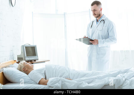Arzt mit Klemmbrett und kranken älteren Frau liegend auf Bett Stockfoto