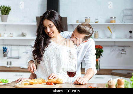 Schöner Mann schneiden Brot auf Schneidebrett und küssen Freundin Schulter Stockfoto