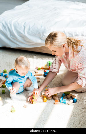 Attraktive blonde Frau sitzt auf dem Teppich in der Nähe von toddlersohn spielen mit Bauklötzen Stockfoto