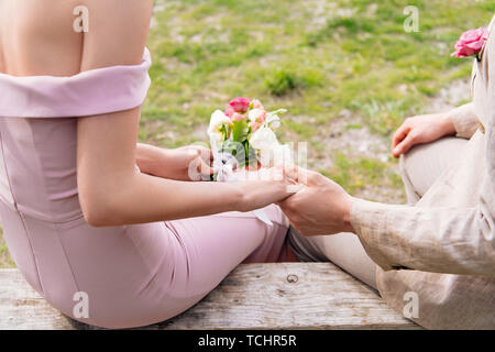 7/8-Ansicht der Braut mit Blumenstrauß und Bräutigam sitzt auf der Bank und halten sich an den Händen Stockfoto