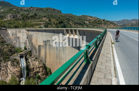 Weibliche Radfahrer reiten über den Damm am Stausee Embalse de Béznar mit dem Wasserfall, Presa de Béznar, im Hintergrund, in der Sierra Nevada, Spai Stockfoto