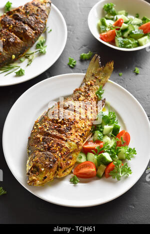 Gekochter Fisch mit gesunden Salat von frischem Gemüse auf dem Teller über schwarzen Stein. Stockfoto