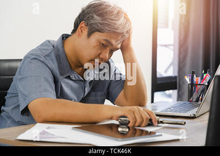 Mann denkt das Arbeiten im Home Office. Sitzt von der Arbeit gelöscht, die linke Hand hält den Kopf wegen der Kopfschmerzen. Mit der rechten Hand eine Pistole ist Stockfoto