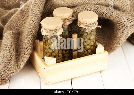 Gläser mit verzinnten Kapern in Sack auf weißem Holz- Hintergrund Stockfoto