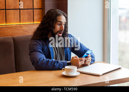 Portrait von schockiert stattlichen Intelligenz junger erwachsener Mann Freiberufler in lässigen Stil im Cafe holding Telefon sitzend mit entsetzten Gesicht, bussinessman in o Stockfoto