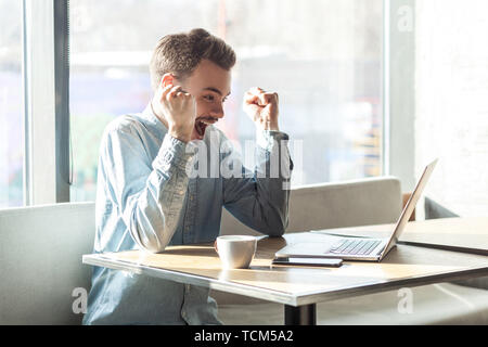 Portrait von Stattlichen erstaunt zufrieden positive bärtige junge Freiberufler in blaues Hemd sitzen im Café und beobachten online Fußballspiel mit toot Stockfoto