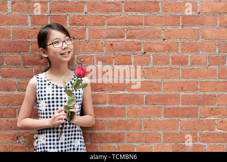 Portrait von jungen asiatischen Mädchen mit roten Rose Blume gegen Red brick wall Stockfoto