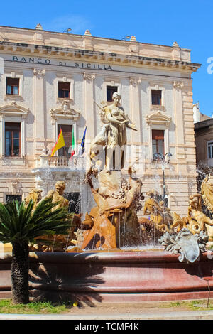 Syrakus, Sizilien, Italien - Apr 10 2019: Erstaunliche Brunnen der Diana auf dem Archimedes Platz im schönen Insel Ortigia. Aufbau einer sizilianischen Bank im Hintergrund. Sonnigen Tag, blauer Himmel. Stockfoto