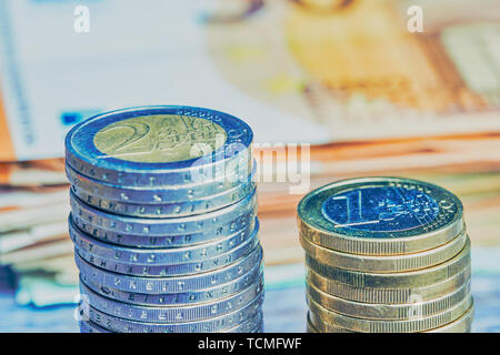 Mit einem Euro Münzen Stapel neben einem von zwei Euro Münzen stack, im Hintergrund liegende verschwommen Euro-banknoten, kalte farbe Stil Stockfoto