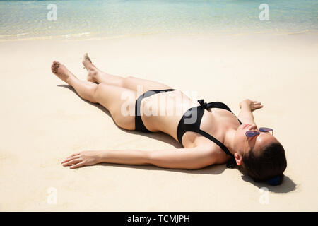 Junge Frau im schwarzen Bikini, Sonnenbrille, entspannend am Sandstrand in der Nähe des Meeres Stockfoto