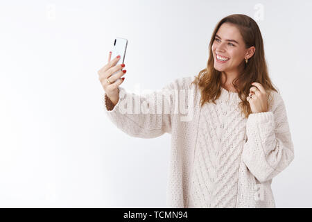 Freudige charismatische zarte weibliche europäische Frau mit gemütlichen Pullover posing Neigekopf cute flirty exted Arm holding Smartphone unter selfie Maki Stockfoto