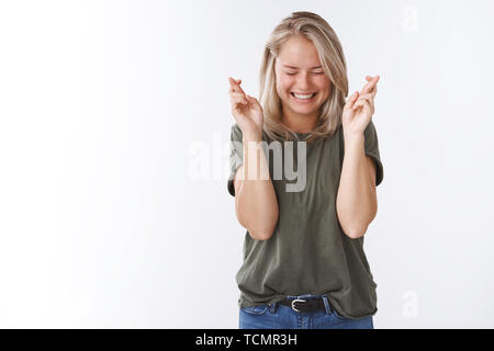 Hoffen auf gute Nachrichten. Portrait von gut - aufgeregt, blonde Frau in Olive t-shirt schließen Augen begeistert, amüsiert lächelnd und halten die Daumen für gekreuzt Stockfoto